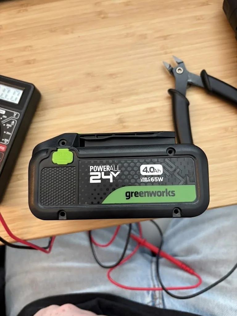 A 24V 4.0Ah Greenworks cordless tool battery sitting on a workbench next to a multimeter and flush cutters.