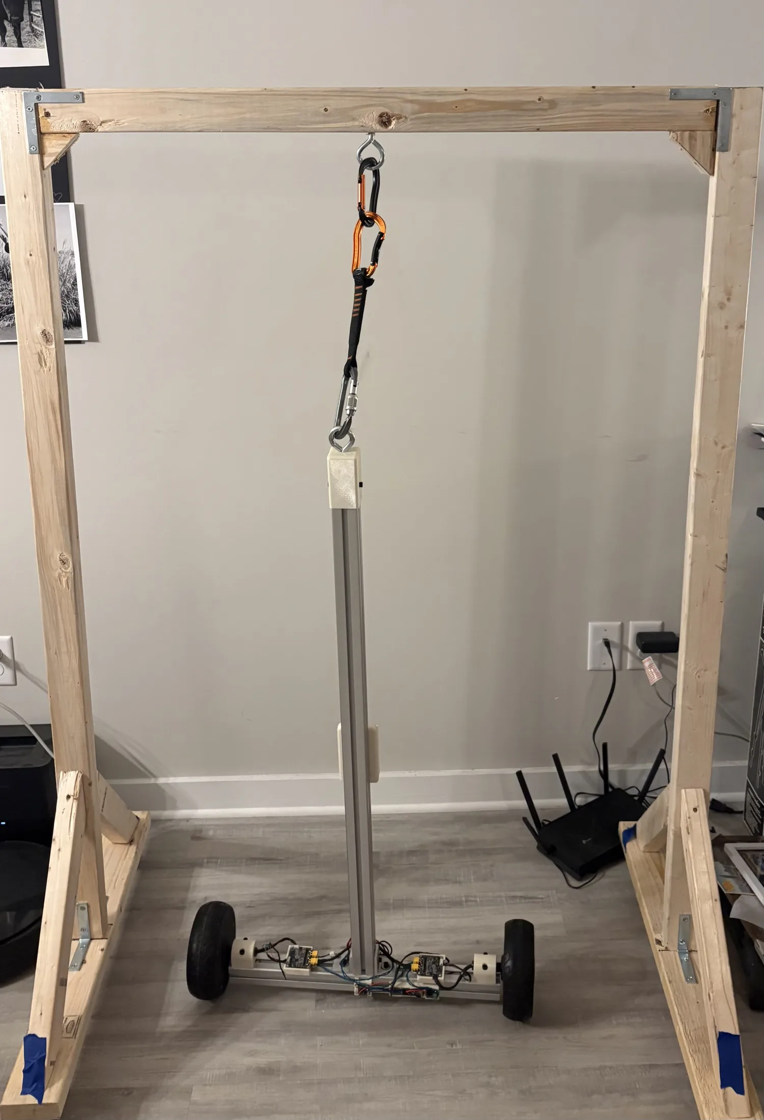 A two-wheeled robot frame made of aluminium extrusion, hanging from a carabiner under a wooden test gantry in an apartment.